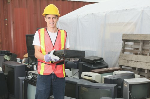 A young male worker wearing a yellow safety helmet and a high-visibility vest with orange and yellow reflective stripes is smiling while holding a black electronic device, possibly a tablet or scanner, in an outdoor setting. The background includes a makeshift outdoor area with a red corrugated metal fence, a white canopy or tent, and a wooden pallet leaning against the fence. On the ground, there are multiple old, discarded computer monitors and electronic waste stacked closely together, indicating a site related to electronic rubbish collection or disposal. The environment suggests a designated space for rubbish removal, with a focus on electronic waste, and the scene underscores the importance of proper hazardous waste handling, as managed by professional rubbish removal services such as Garden Clearance Westminster servicing Westminster and surrounding areas.